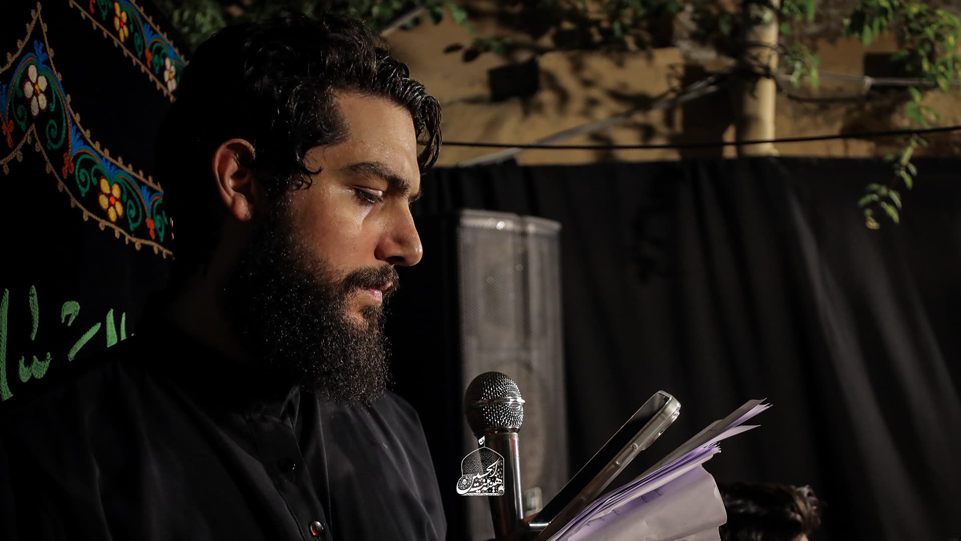 A reciter reading elegies at the microphone beneath embroidered black banners during a majlis