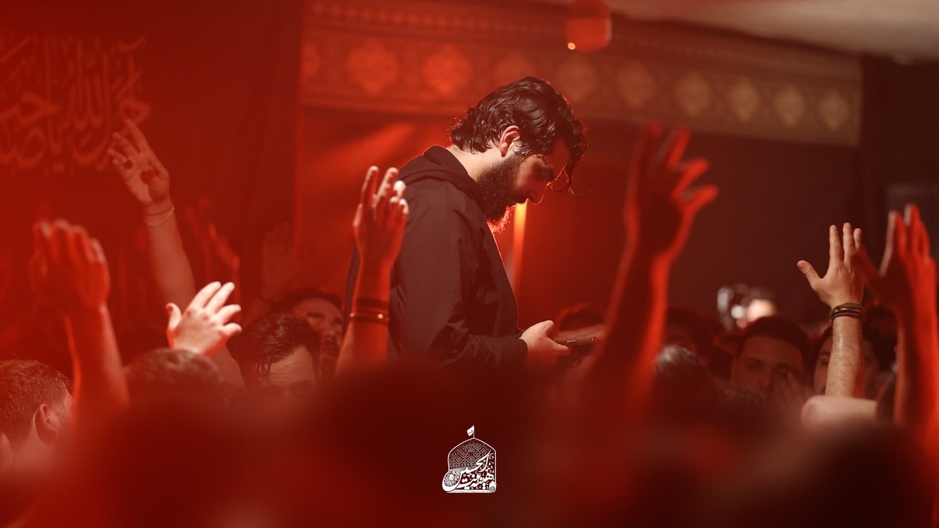 A reciter standing above the crowd in deep red light as mourners raise their hands